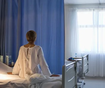 A patient in a hospital gown sits on a bed facing a blue curtain in a sunlit room.