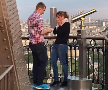 Man proposing to a woman at the top of the Eiffel Tower, with a scenic city view in the background.