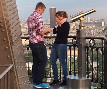 Man proposing to a woman at the top of the Eiffel Tower, with a scenic city view in the background.