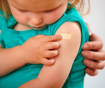 Child wearing a turquoise shirt showing a bandage on their arm, with an adult hand gently supporting them.