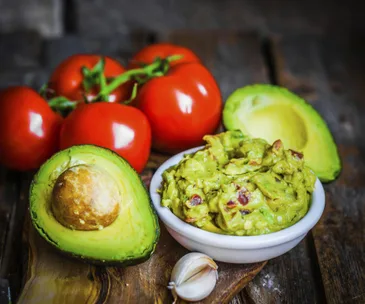 Bowl of guacamole with tomatoes, avocados, and garlic on a wooden surface.