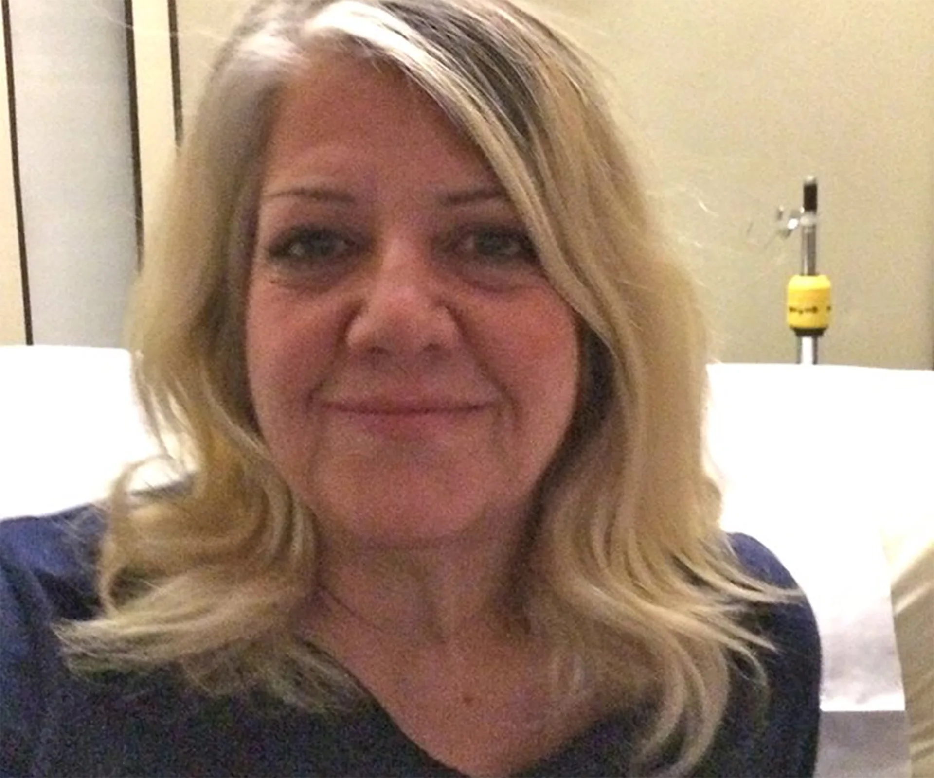 Smiling woman with blonde hair in a room, seated in front of a blurred background.