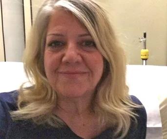 Smiling woman with blonde hair in a room, seated in front of a blurred background.