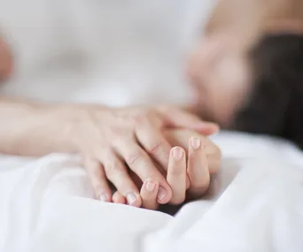 Close-up of two hands gently holding on a white bed, with a blurred background.