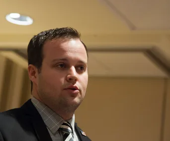 Man speaking in a dimly lit room, wearing a suit and tie.