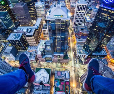 Aerial view of Melbourne cityscape at dusk, showing skyscrapers and streets with person's feet dangling from a building ledge.