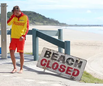 Lifeguard in red and yellow uniform near "Beach Closed" sign with empty beach in background.
