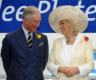 A man in a blue suit and a woman in a cream outfit with a large hat smiling at each other outdoors.