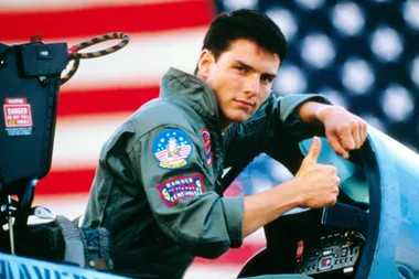 Pilot in cockpit giving thumbs up, with American flag in background.
