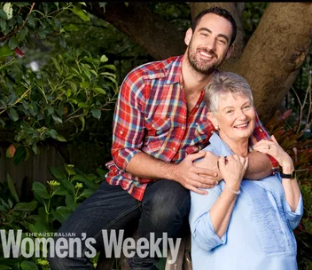 Man in plaid shirt hugs smiling woman in blue blouse in a garden setting, with The Australian Women's Weekly logo.