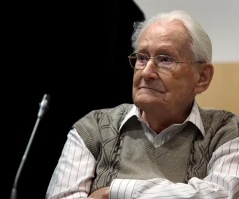 An elderly man with glasses sits solemnly in a courtroom setting, wearing a striped shirt and patterned sweater.