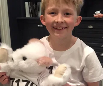 Young boy smiling while holding a white stuffed animal in a room with dark cabinets.