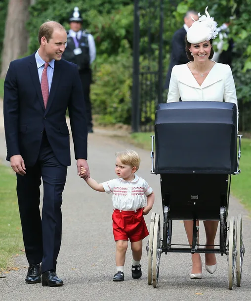 Royal family walking outdoors; a man, child, and woman pushing a vintage pram.