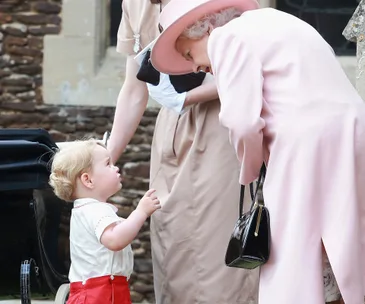 A young child looks up at an elderly woman dressed in pink, carrying a black purse, outside near a stone building.