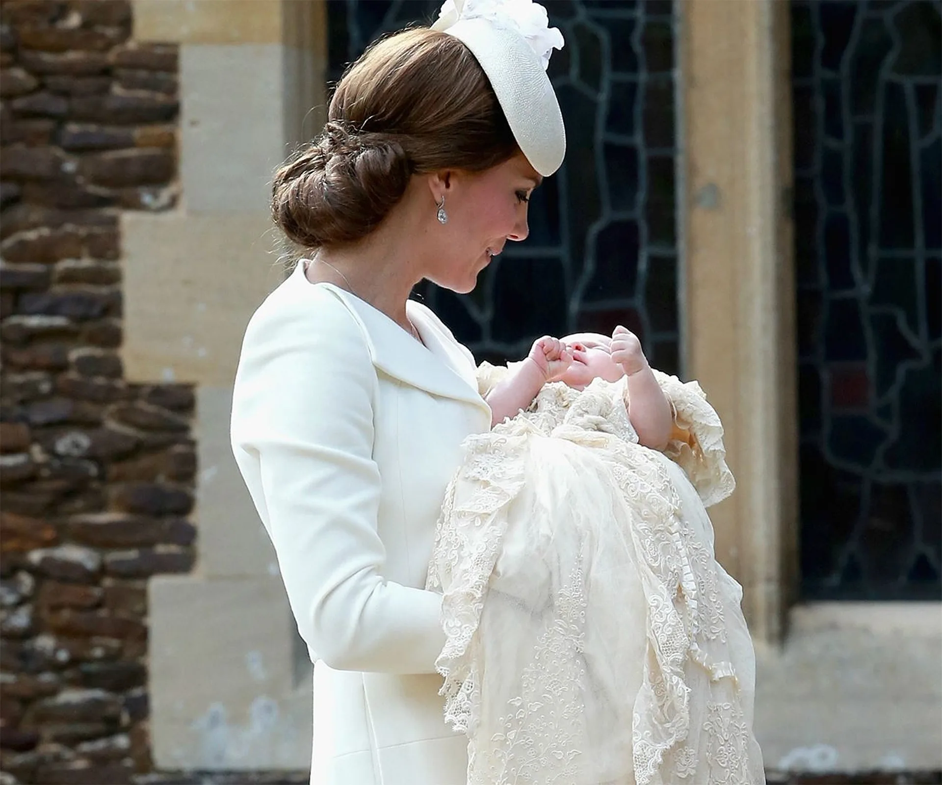 A woman in a white outfit holding a baby in a lace christening gown outside a building.