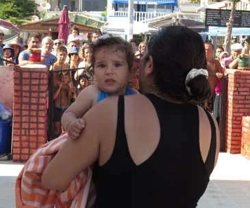 A woman holds a baby wrapped in a towel, with a crowd watching behind a brick wall.