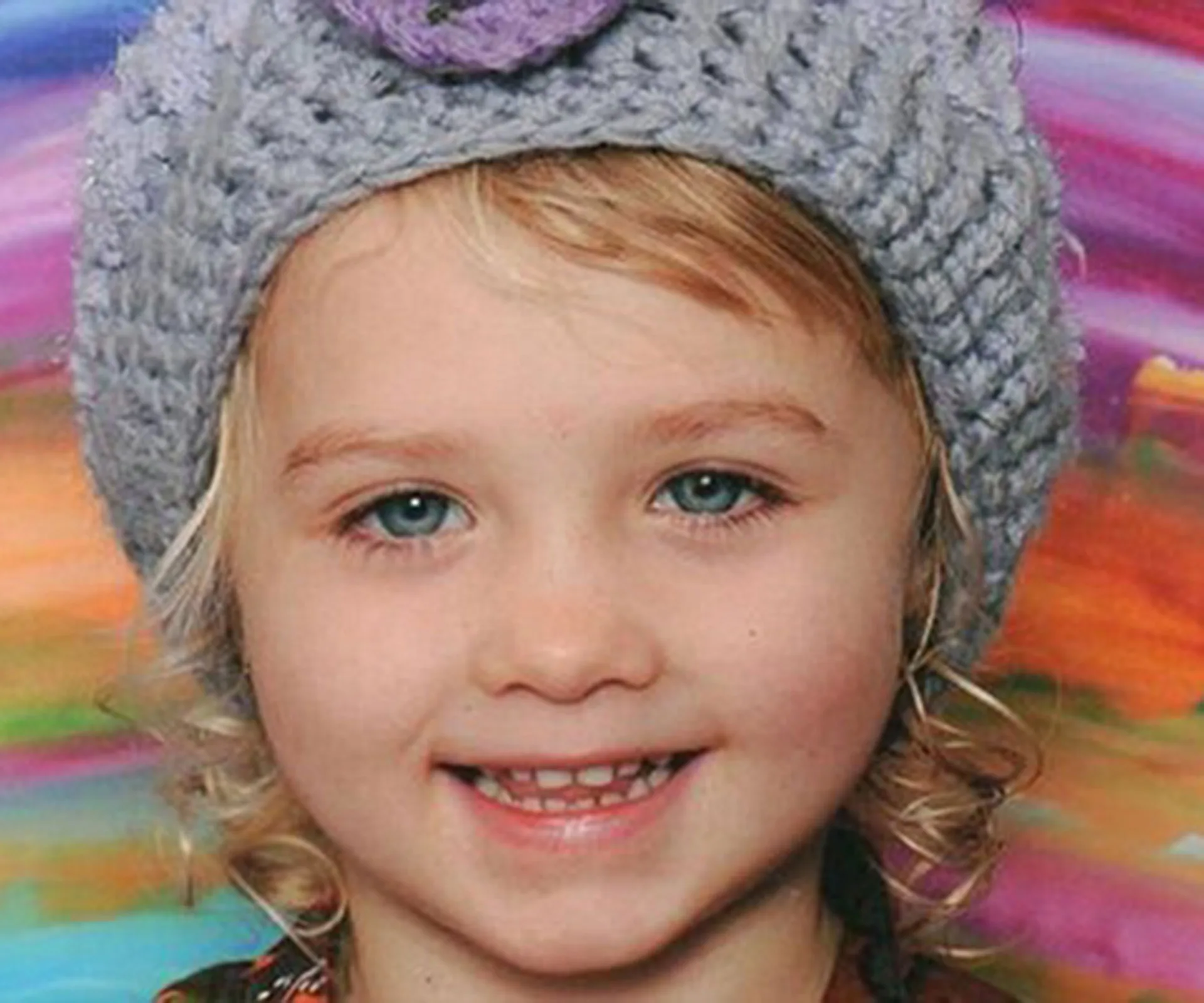 A young girl with curly blonde hair, wearing a knitted hat, smiles in front of a colorful background.