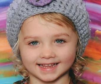 A young girl with curly blonde hair, wearing a knitted hat, smiles in front of a colorful background.