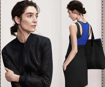 Model in perforated black jacket and black dress with blue top, carrying a black tote bag.