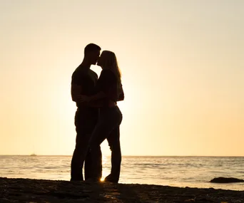 A silhouetted couple stands intimately on a beach at sunset, with a sailboat in the background.