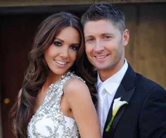 A couple smiling at the camera, woman in an embellished dress, man in a suit with a white lily boutonniere.