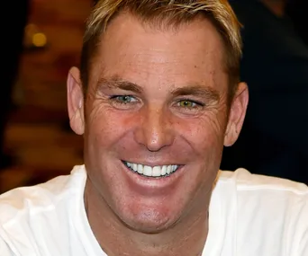 A person smiling broadly, wearing a white shirt, against a blurred background.