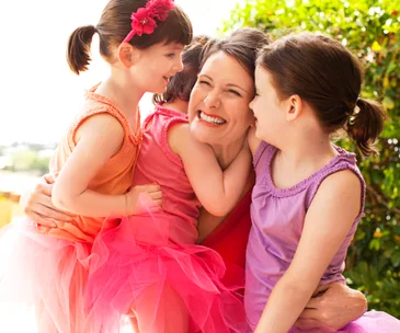 A smiling mother embraces her two young daughters wearing colorful dresses and tutus in a sunny outdoor setting.