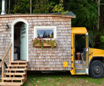 Yellow school bus converted into a tiny home with wooden siding, flowers in window box, and wooden steps leading to a door.