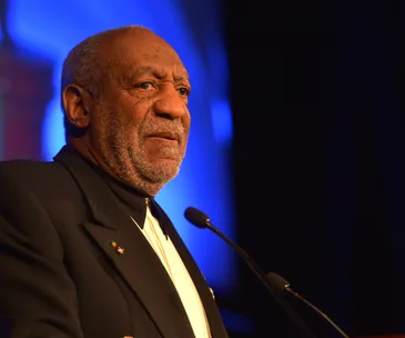 A man in a suit speaks at a podium, lit with blue lighting in the background.
