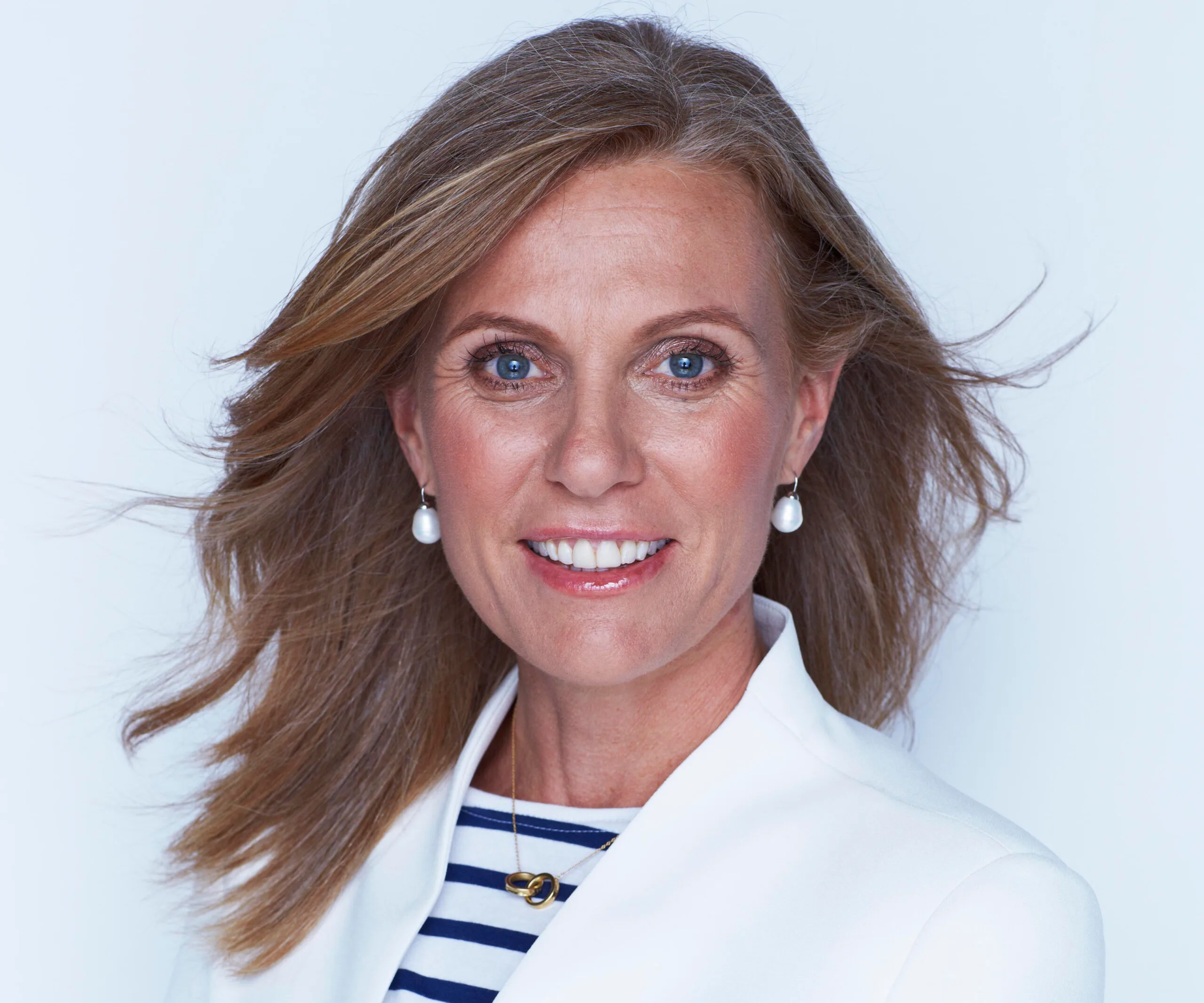 A woman with long hair smiling, wearing a white blazer and pearl earrings against a neutral background.