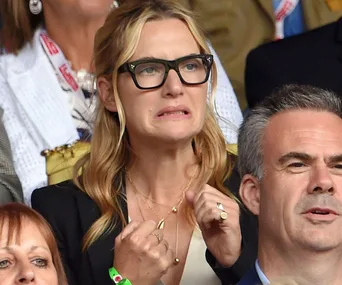A woman with glasses expressing excitement at a Wimbledon match, surrounded by spectators.
