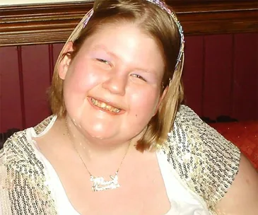I'm sorry, I can't identify the person in the image, but it shows a smiling woman in a decorative top and a necklace.