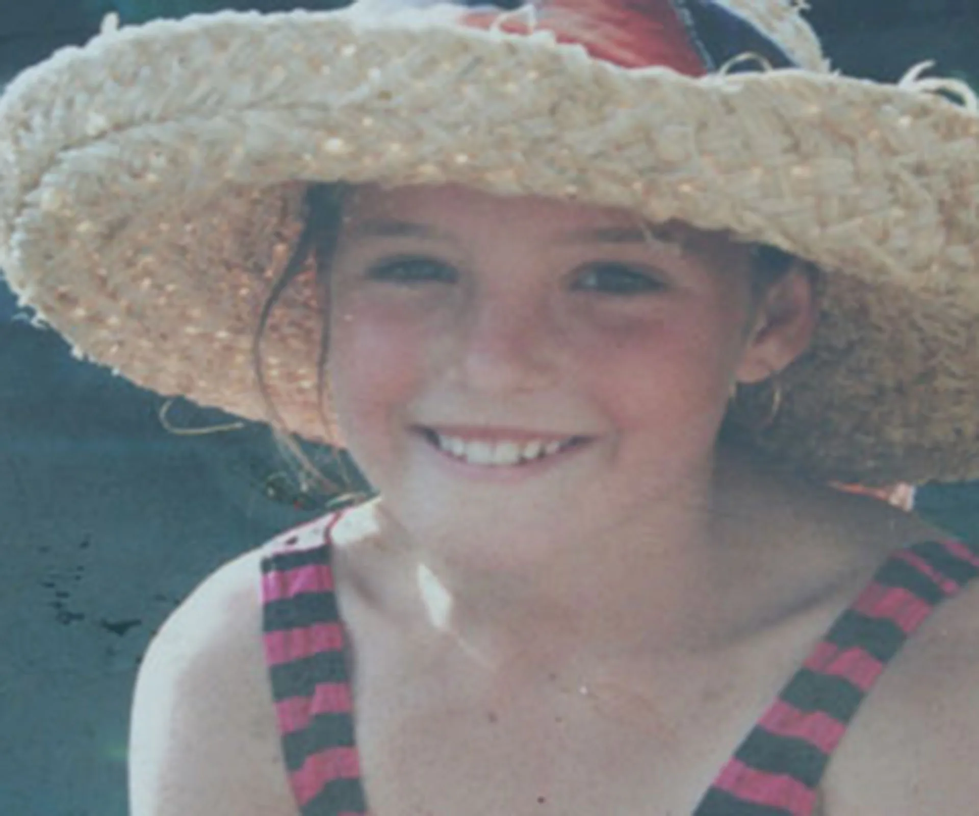 A young girl smiling, wearing a sun hat and a striped swimsuit, outdoors on a sunny day.