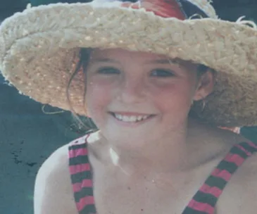 A young girl smiling, wearing a sun hat and a striped swimsuit, outdoors on a sunny day.