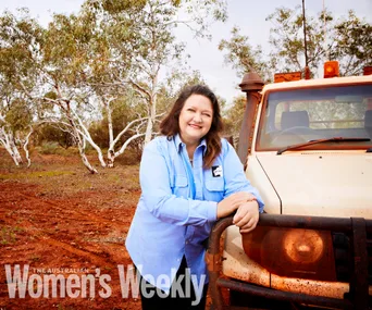 Woman in blue shirt leans on a 4WD in an Australian outback setting, with gum trees in the background.
