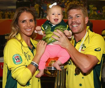 Australian cricketer and partner in yellow jerseys holding their baby on ICC trophy.