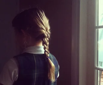 A child with a braided hairstyle stands by a window in school uniform, facing away from the camera.