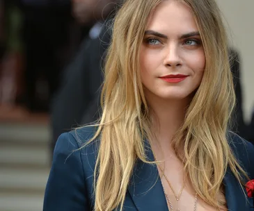 A woman with long blonde hair and red lipstick, wearing a dark blazer, smiles softly, looking to the side.