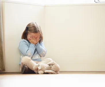 Child in blue sweater sits against a wall, covering face with hands, teddy bear on floor.