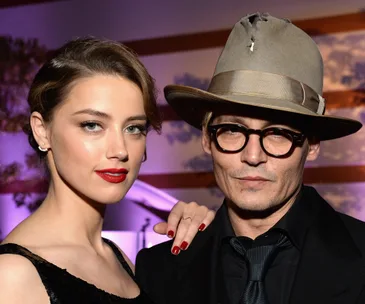 Two individuals posing closely; one with red lipstick and the other in glasses and a hat, in a formal setting.