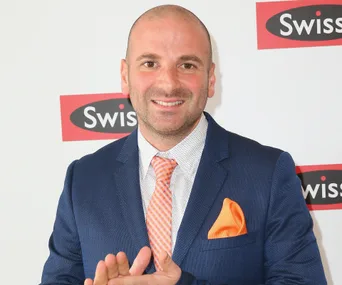 Man in a blue suit with orange tie and pocket square, standing in front of a Swisse logo backdrop.