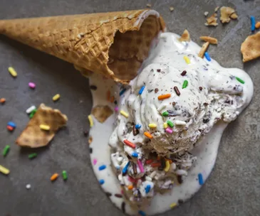 Melted ice cream with sprinkles spilling from a fallen waffle cone on a gray surface.