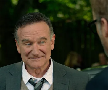 ```plaintext
A man in a suit smiling slightly while outdoors, with blurred greenery in the background.
```