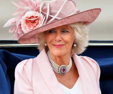 A woman in a pink hat with a large flower, pearl necklace, and light-colored coat, smiling while seated outdoors.