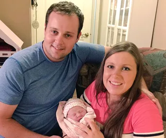 A couple smiles while holding their newborn baby, wrapped in a patterned blanket.