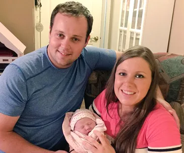 A couple smiles while holding their newborn baby, wrapped in a patterned blanket.