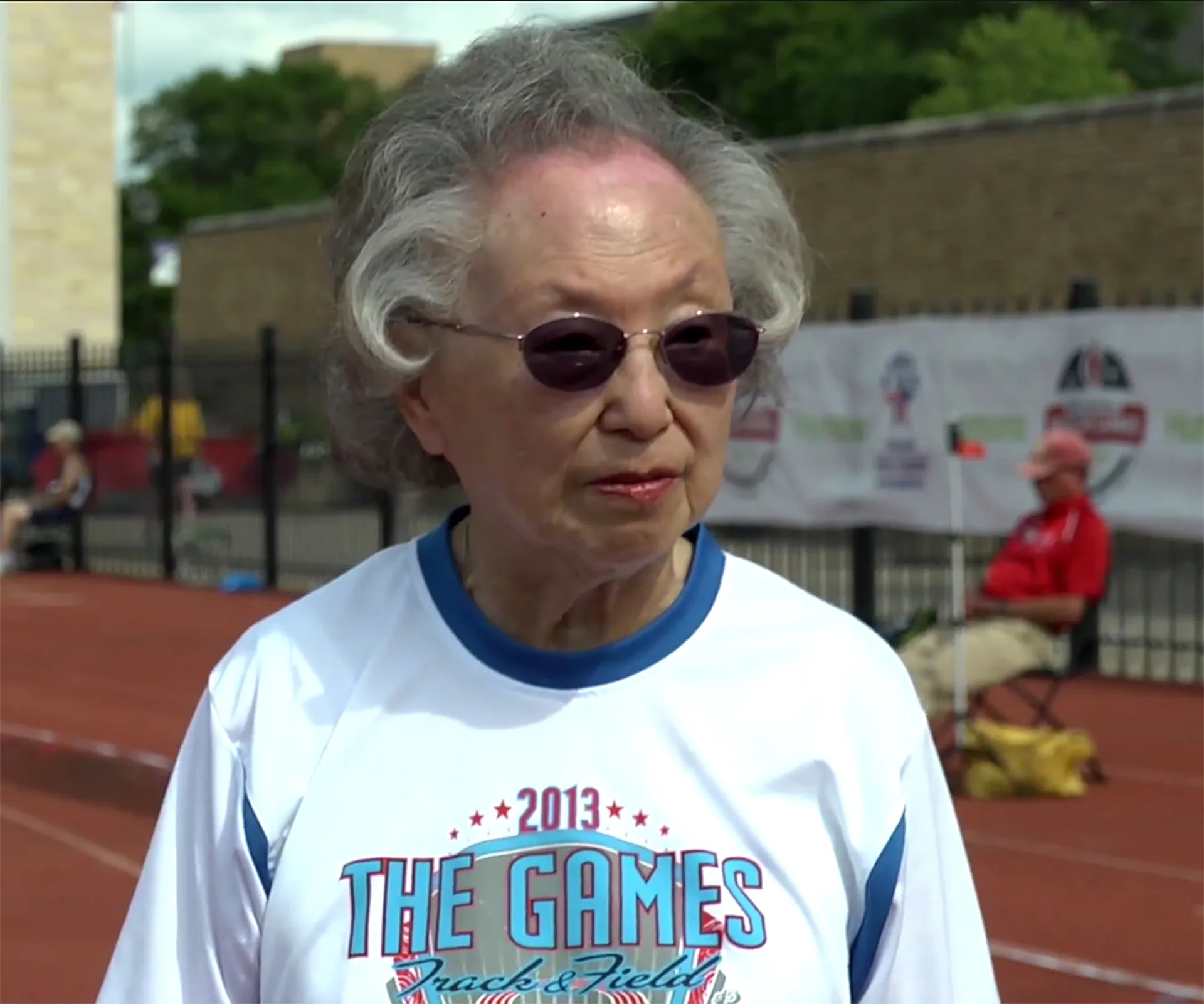 Elderly woman in sunglasses wearing "2013 The Games Track & Field" shirt at a sports event.