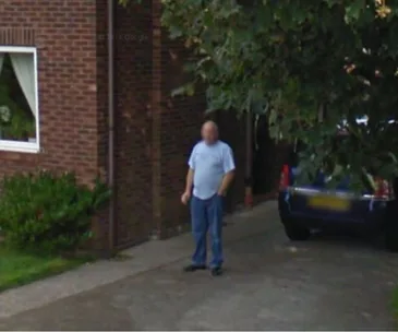 A man stands on a driveway next to a parked car in front of a brick house.