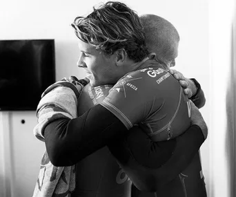 Two surfers embrace warmly, each wearing surf gear, conveying a sense of camaraderie and support.