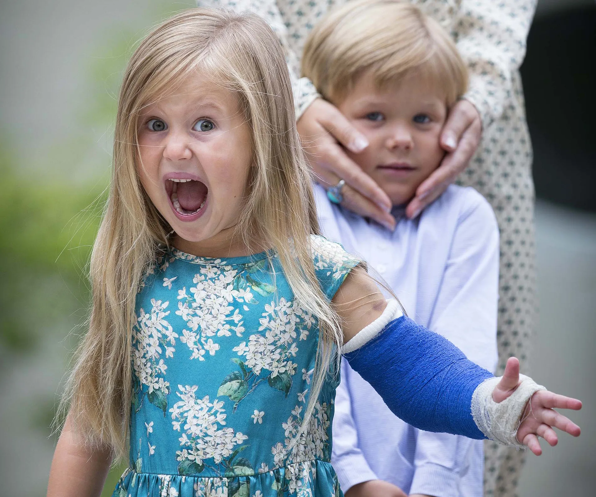 Girl in floral dress with blue cast, mouth open in excitement; boy behind her looking shy, hands on his face.